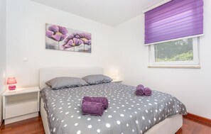 Un dormitorio con cama, mesita de noche, lámpara, ventana con persianas y un cuadro en la pared.