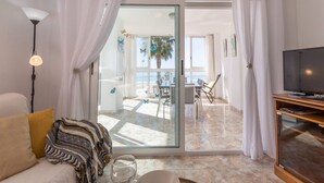 A modern living room with a sofa, a glass door leading to a balcony with a view of the sea, and a television on a wooden stand.