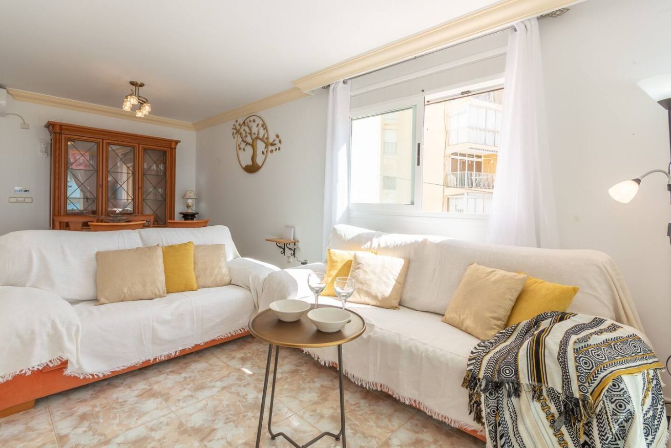 A living room with a white sofa, a small round table with bowls, a patterned throw blanket, and a window with a view of a building.