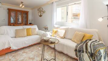 A living room with a white sofa, a small round table with bowls, a patterned throw blanket, and a window with a view of a building.