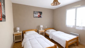 Een kamer met twee houten bedden, wit beddengoed, een aan de muur hangend schilderij, een hanglamp en een raam met dunne gordijnen.