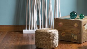 Une pièce dotée d’un plancher en bois, d’un pouf en osier, d’une colonne décorative blanche et d’une malle en bois ornée d’autocollants.