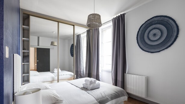 Une chambre moderne avec un grand miroir, un lit recouvert de draps blancs, un luminaire suspendu et une œuvre d’art circulaire fixée au mur.