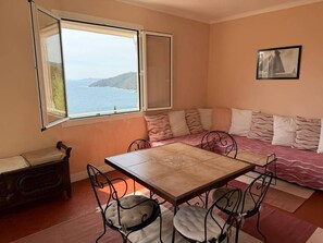 Une pièce chaleureuse avec une table à manger, des chaises, un canapé avec des coussins, une fenêtre donnant sur la mer et un tableau encadré au mur.
