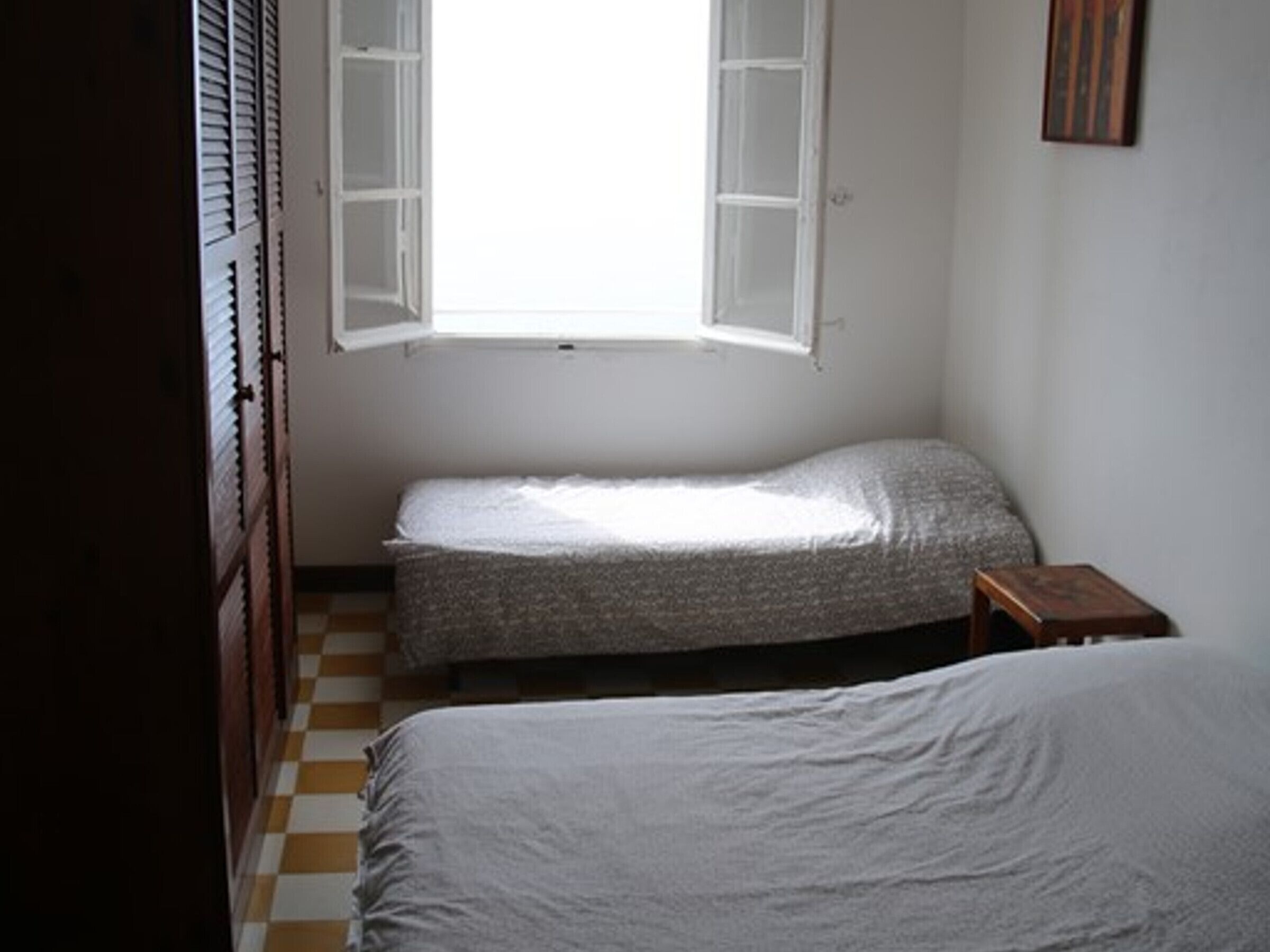 A small, dimly lit room with two single beds, a wooden wardrobe, and a window with open shutters.