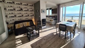 Een moderne woonkamer met een bank, salontafel en een eettafel. Er is een keuken met uitzicht op de zee.