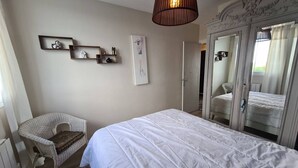 Een slaapkamer met een groot bed, een rieten stoel, een houten dressoir en een spiegel.