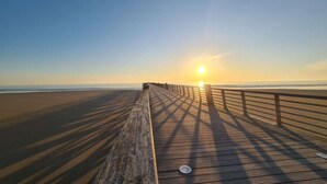 Een houten looppad dat naar het strand leidt, met een heldere lucht en een ondergaande zon in de verte.