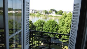 A view from a window with shutters, overlooking a river with boats, greenery, and buildings in the distance.