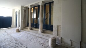 A building entrance with a stone-paved area, columns, and a staircase leading to a dark door.