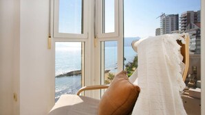 Une chambre dotée d’une grande fenêtre donnant sur la côte, d’un lit recouvert d’une couverture blanche et d’un oreiller marron.