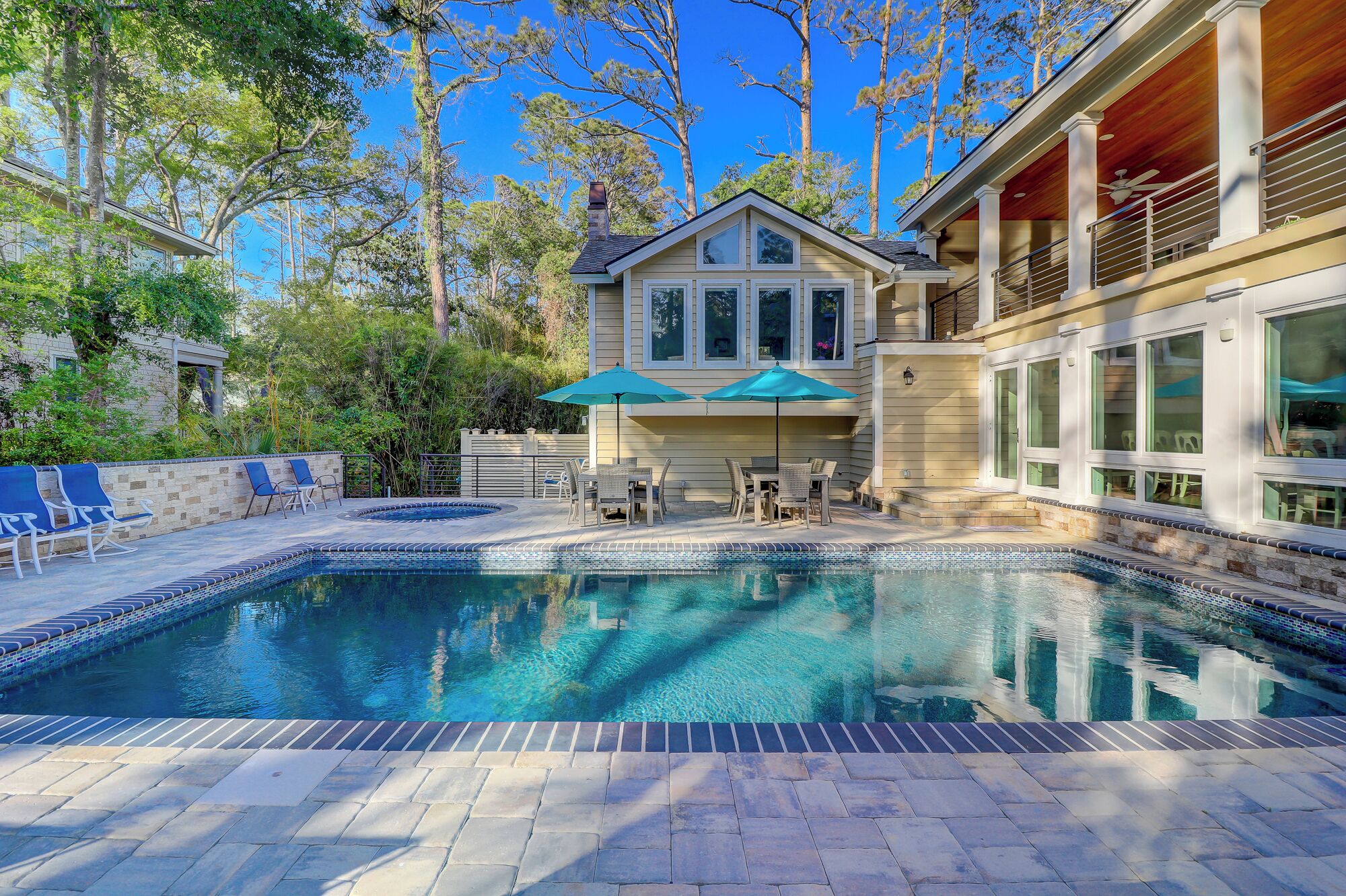 A swimming pool with a tiled deck, a covered seating area with umbrellas, and a house with large windows and a balcony.