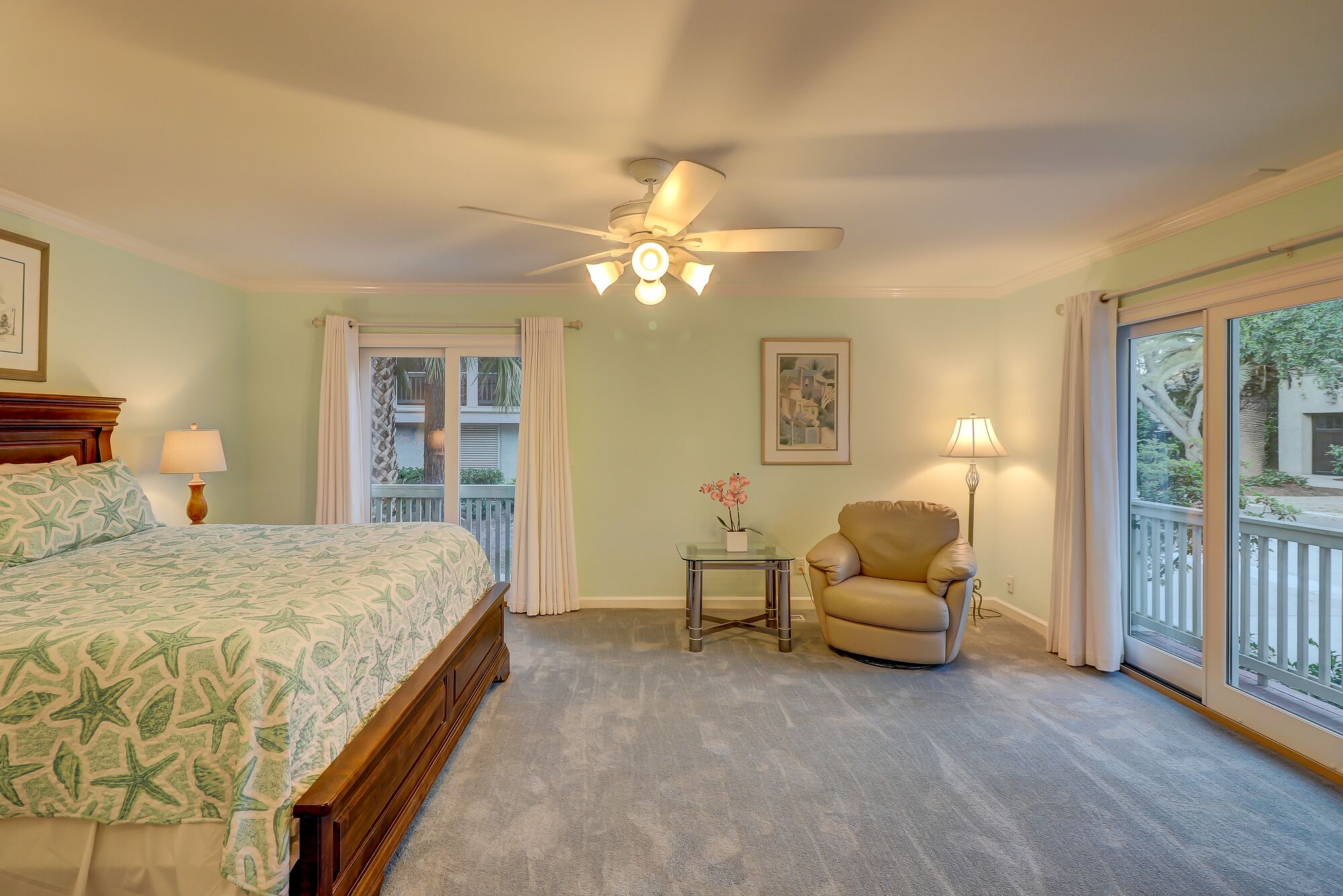 A bedroom with a bed, a chair, a small table, a ceiling fan, and a window with a view of the outside.