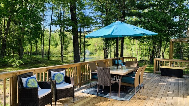 Una terraza de madera con mobiliario exterior, incluyendo sillas de mimbre y una mesa, bajo un paraguas azul.