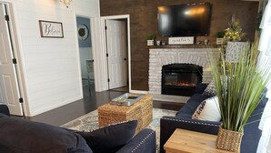 Una sala de estar moderna con chimenea, televisor montado en la pared y un área de descanso confortable con sofá y sillones.