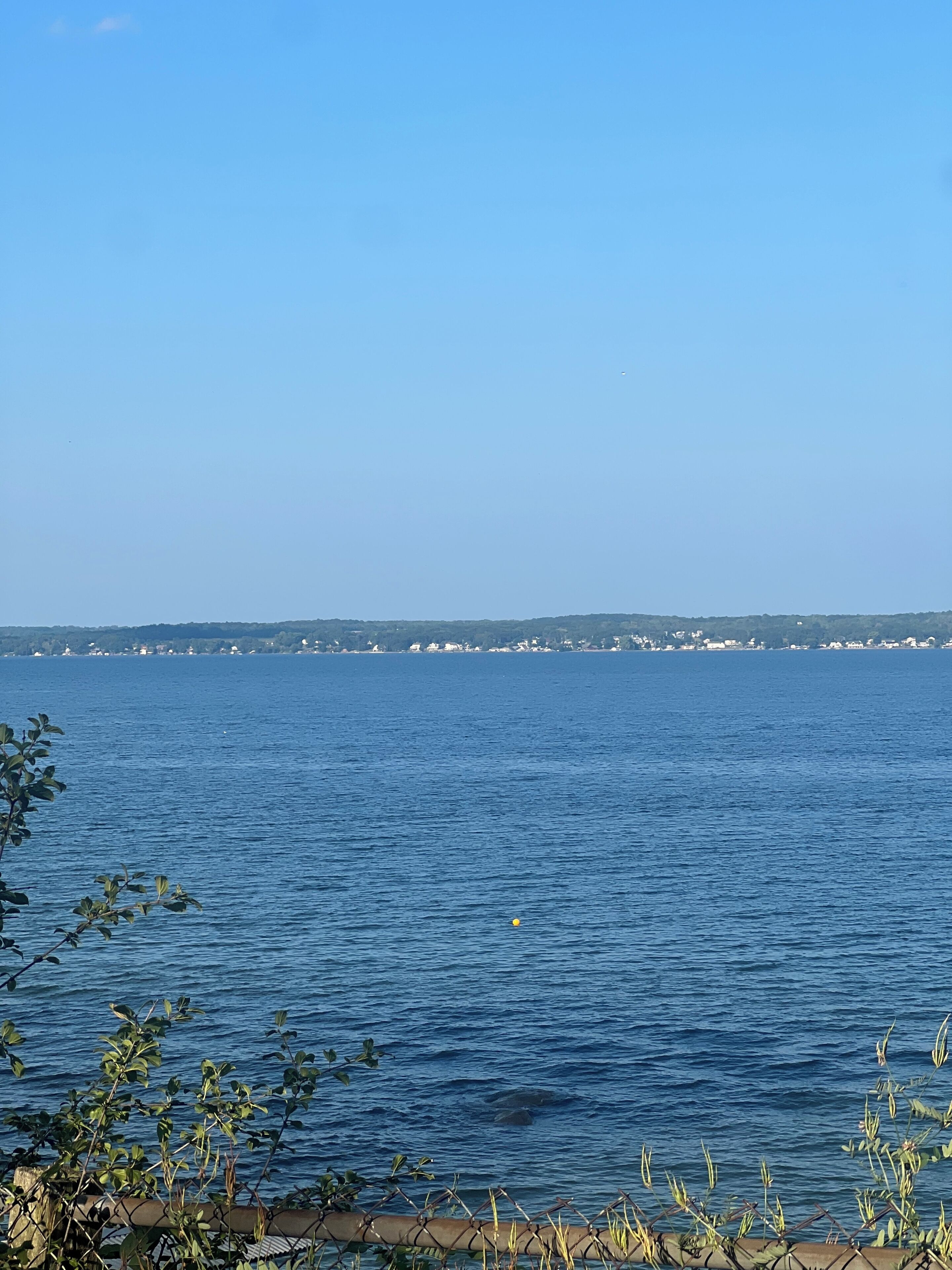 A coastal view with calm waters, a distant shoreline with buildings, and a clear sky.