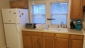 A kitchen with wooden cabinets, a refrigerator, a toaster, a sink, and a window with blinds. - Adorable Cape Cod House in a Little Fishing Town (Marshallberg)