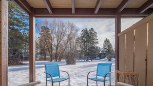 Un espace extérieur aménagé avec une zone de repos couverte, comprenant deux chaises bleues et une table en bois, d’où l’on peut admirer un paysage enneigé avec des arbres et un bâtiment en arrière-plan.