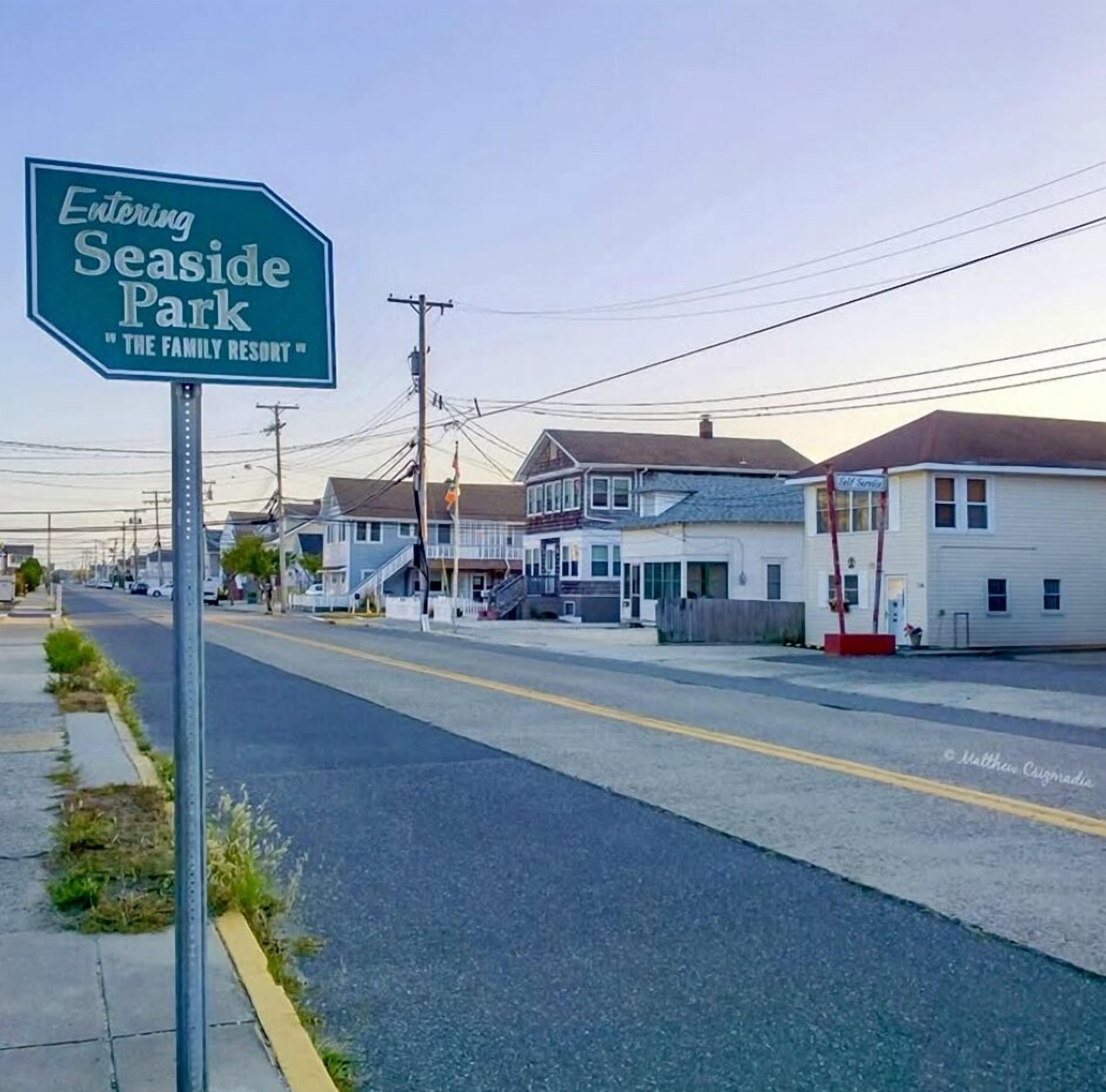 A road sign indicating the entrance to Seaside Park, with residential buildings and a clear sky in the background.