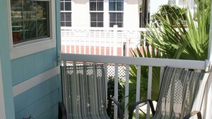 Eine Veranda mit Holzboden, zwei gestreifte Stühle, einem weißen Geländer und Blick auf ein Haus mit weißem Zaun und einer Palme.