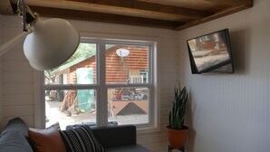 A cozy living room with a gray sofa, a floor lamp, a potted plant, and a large window offering a view of a wooden cabin.