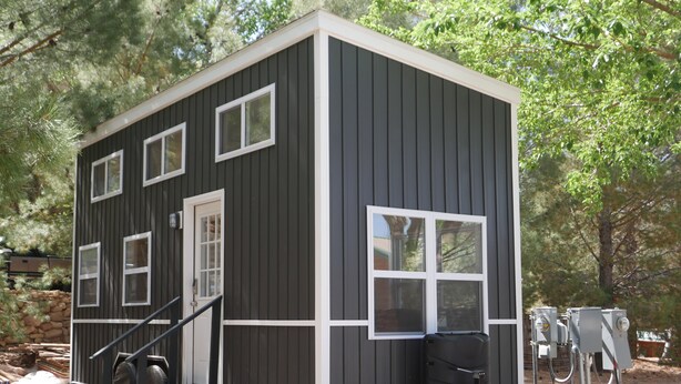 A compact, single-story cabin with a gray exterior and white trim, featuring multiple windows and a small porch area.
