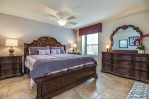A bedroom with a large bed, two bedside tables, a dresser, a ceiling fan, and a window with blinds.