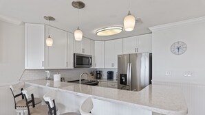 Eine moderne Küche mit weißen Schränken, einem Edelstahl-Kühlschrank, einer Mikrowelle und einem Spülbecken. Drei Pendelleuchten und ein Barhocker mit Sitzkissen.