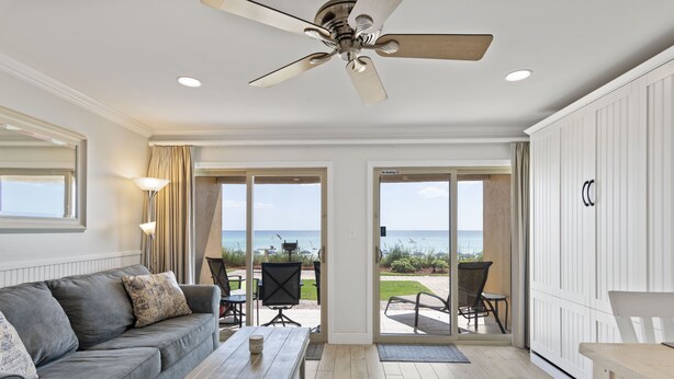 Ein geräumiges Wohnzimmer mit grauem Sofa, einem Holztisch und einem Deckenventilator. Eine Glastür öffnet sich zu einer Außenpatio mit Blick auf den Ozean.
