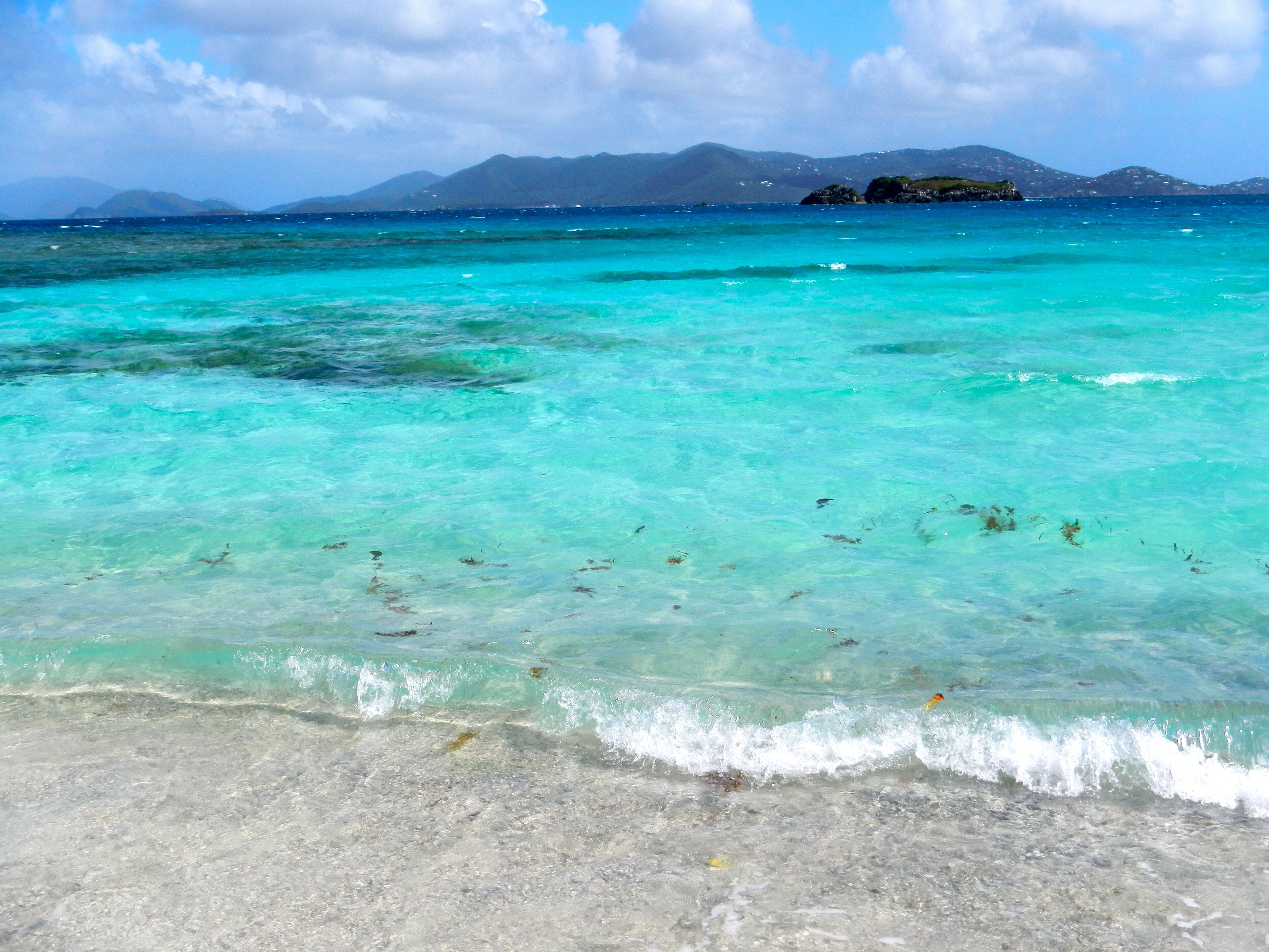 Een strand met helder turquoise water, een zandstrand en op de achtergrond bergen.