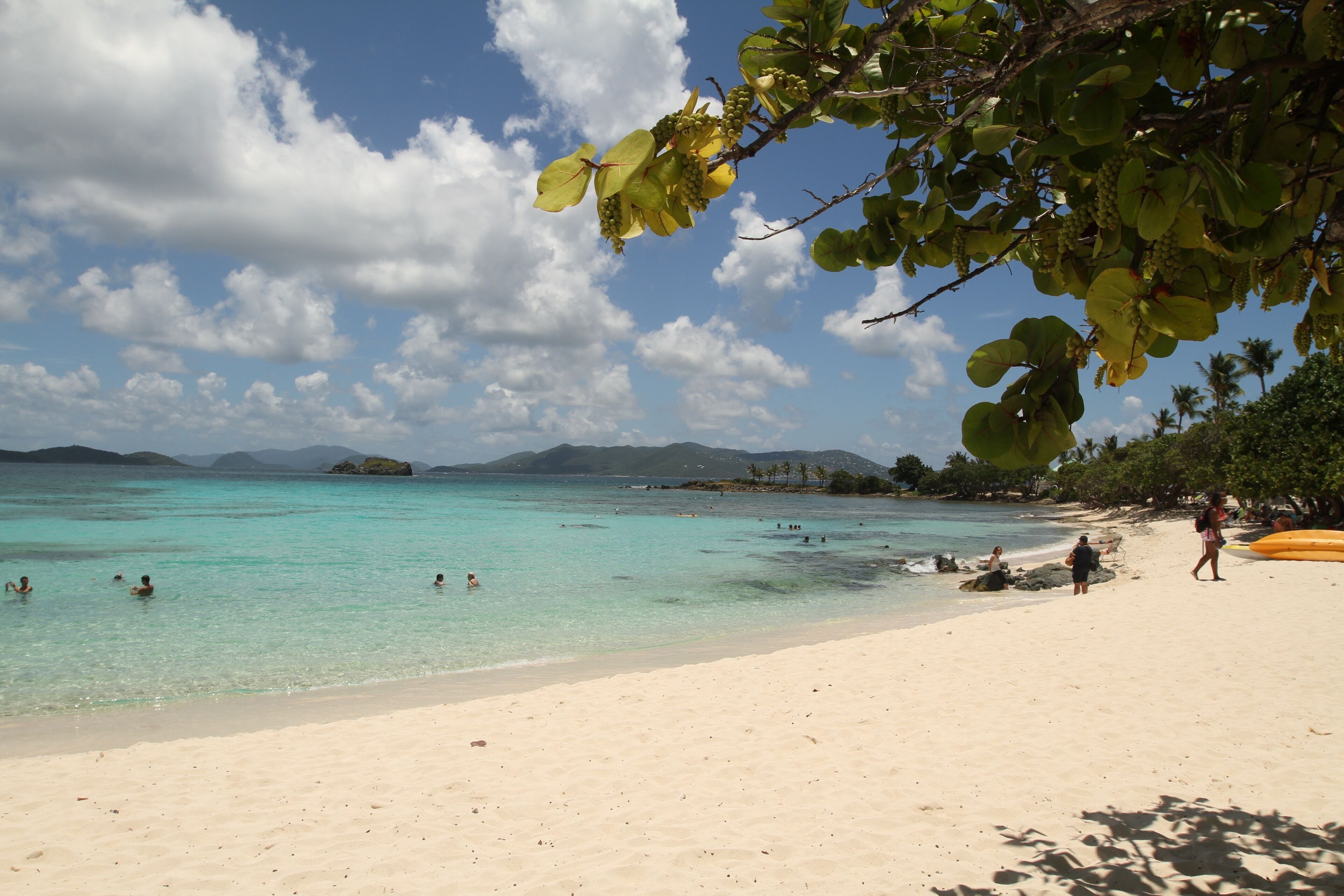 A beach with clear turquoise water, people swimming and walking on the sand, and a clear sky with clouds.