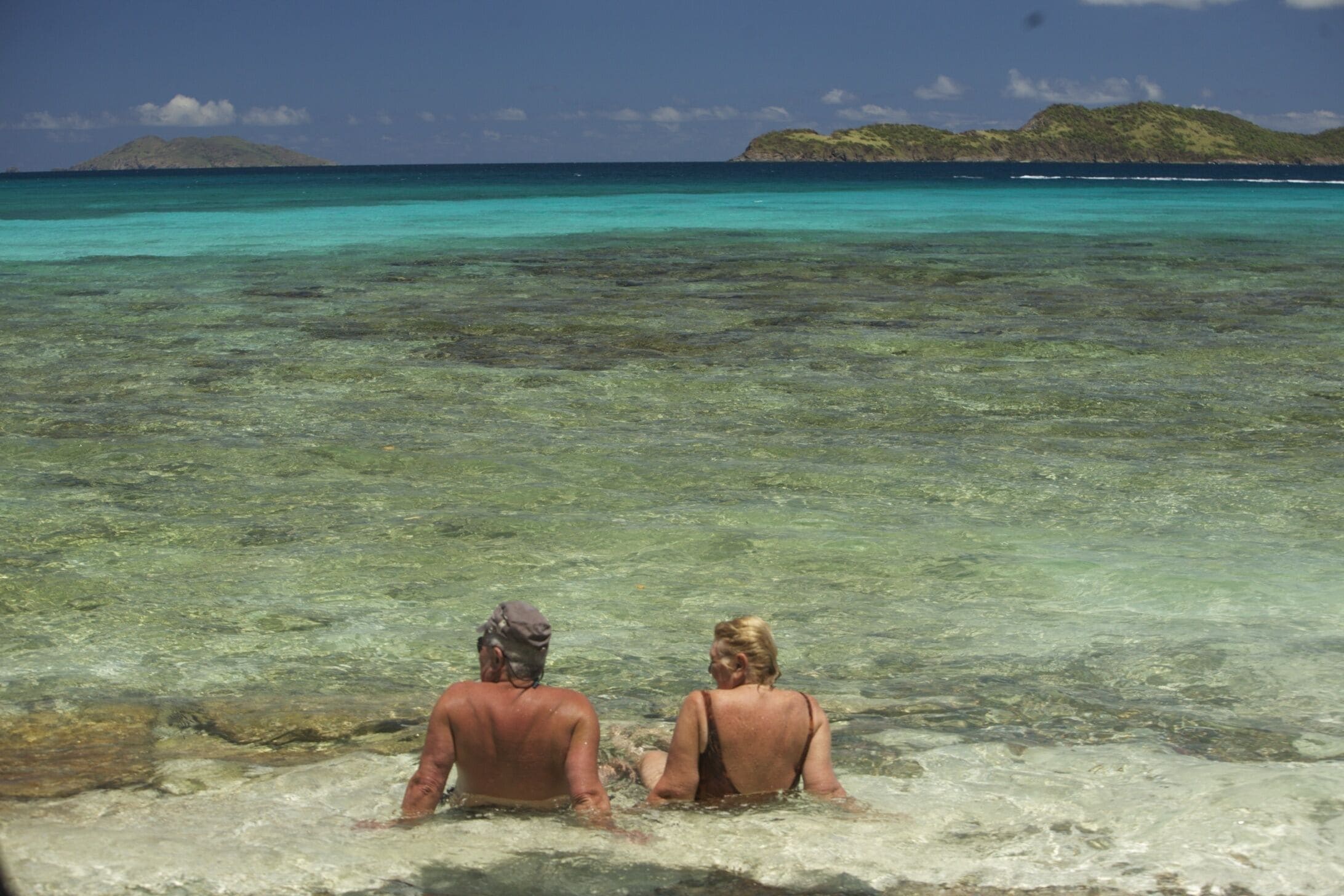 Twee personen zitten in ondiep water, met helder zicht op verre eilanden.
