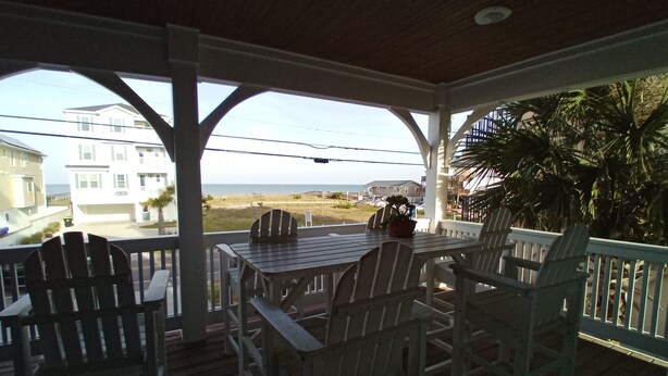A covered outdoor seating area with Adirondack chairs and a table, overlooking a beach and residential buildings.