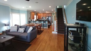 A spacious living room with a sofa, coffee table, and a TV on a stand. A kitchen with wooden cabinets and a dining area with chairs are visible in the background.