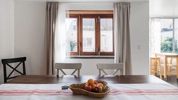 Un comedor con una mesa de madera, dos sillas blancas, una cesta de mimbre para fruta y una ventana con cortinas.