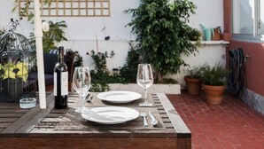 Mesa de madera con un paraguas color beige, una botella de vino y copas, colocados al aire libre en un patio de ladrillo.