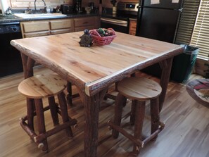 A wooden table with two stools, a basket with pinecones, and a kitchen in the background.