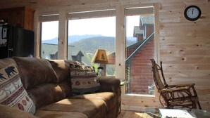 A cozy cabin interior with a leather couch, a wooden chair, a glass table, and a view of mountains through large windows. - Excellent Mountain View’s, Hot Tub, WIFI, Minutes to Gatlinburg (Gatlinburg)