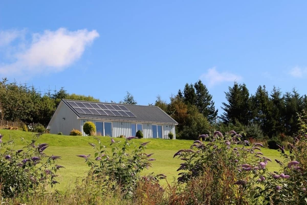 A single-story building with a sloped roof, solar panels, and a small garden area in front.