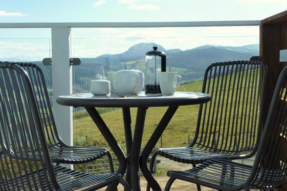 A balcony with a table set for two, featuring a coffee maker, teapot, and cups. The view includes a grassy landscape and mountains in the distance.