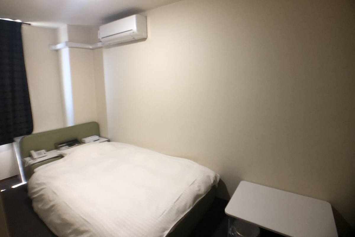 A hotel room with a bed, a bedside table, a phone, and an air conditioning unit.