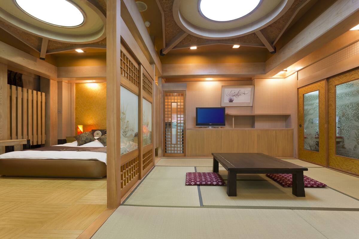 A traditional Japanese-style room with tatami flooring, a low wooden bed, a tatami mat table, and a television on a wooden stand.