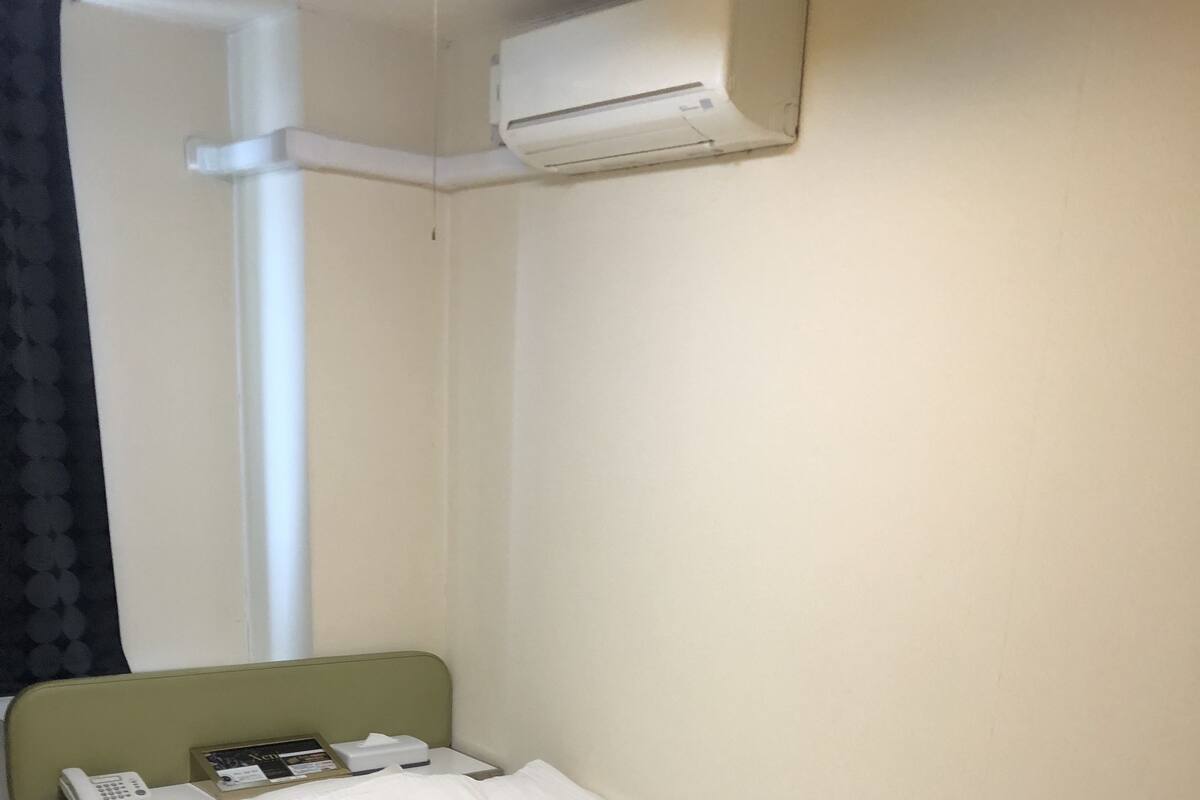 A single bed with a green headboard, a bedside telephone, and a small table with a trash can.