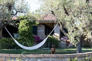 A hammock tied between two trees in a garden area with a stone building in the background.