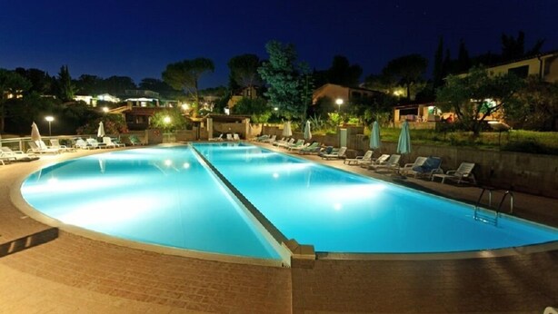A large indoor swimming pool with multiple lanes, surrounded by lounge chairs and umbrellas, set against a backdrop of residential buildings and trees. - Il Borgo Centro Vacanze (Guardistallo)