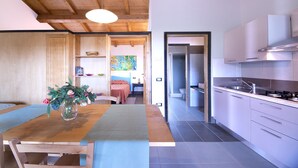 A modern kitchen with wooden beams, a dining area with a table and chairs, and a view into a bedroom with a bed and a colorful painting. - Il Borgo Centro Vacanze (Guardistallo)