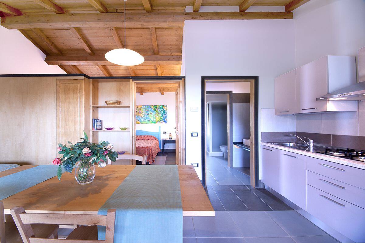 Eine moderne Küche mit Holzbalken, ein Essbereich mit Tisch und Stühlen und ein Blick in ein Schlafzimmer mit Bett und einem farbenfrohen Gemälde.