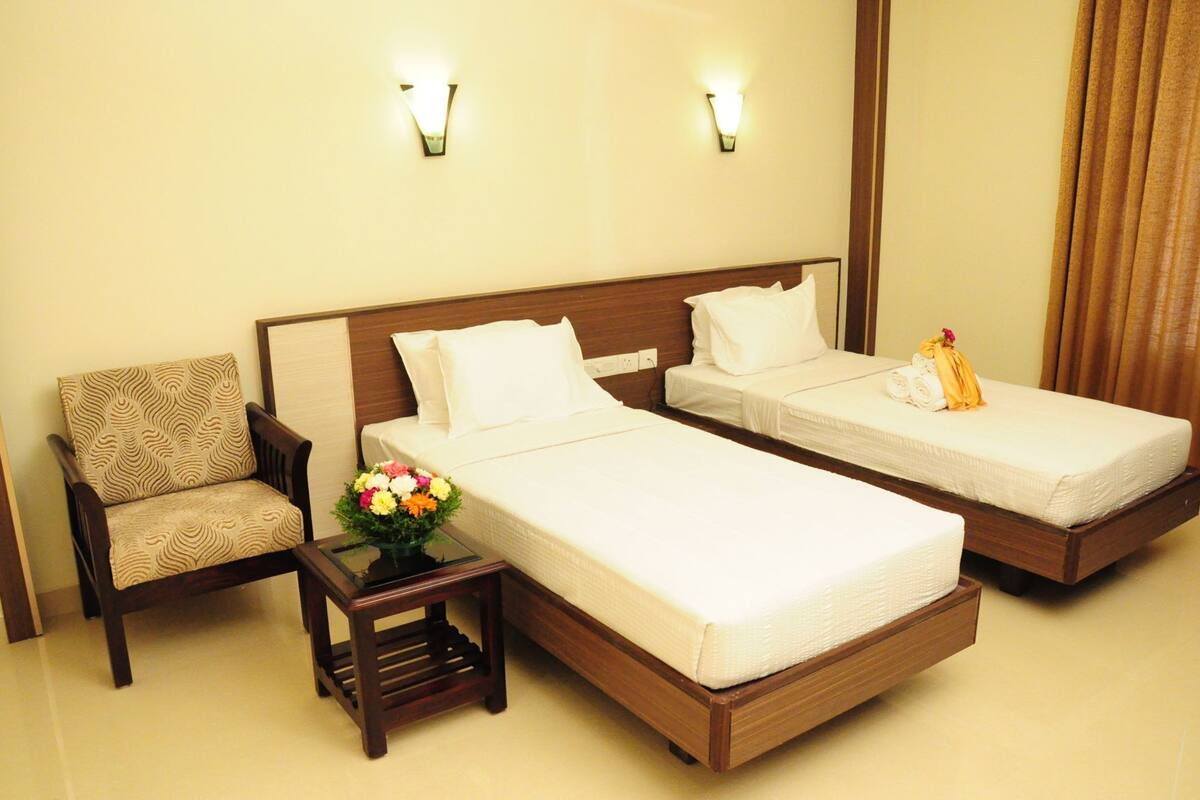 A hotel room with two single beds, a chair, a small table with a vase of flowers, and two wall-mounted lights.