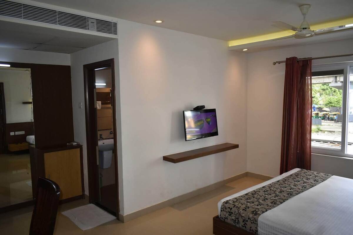 A hotel room with a bed, a television mounted on the wall, a window with curtains, and a door leading to a bathroom.