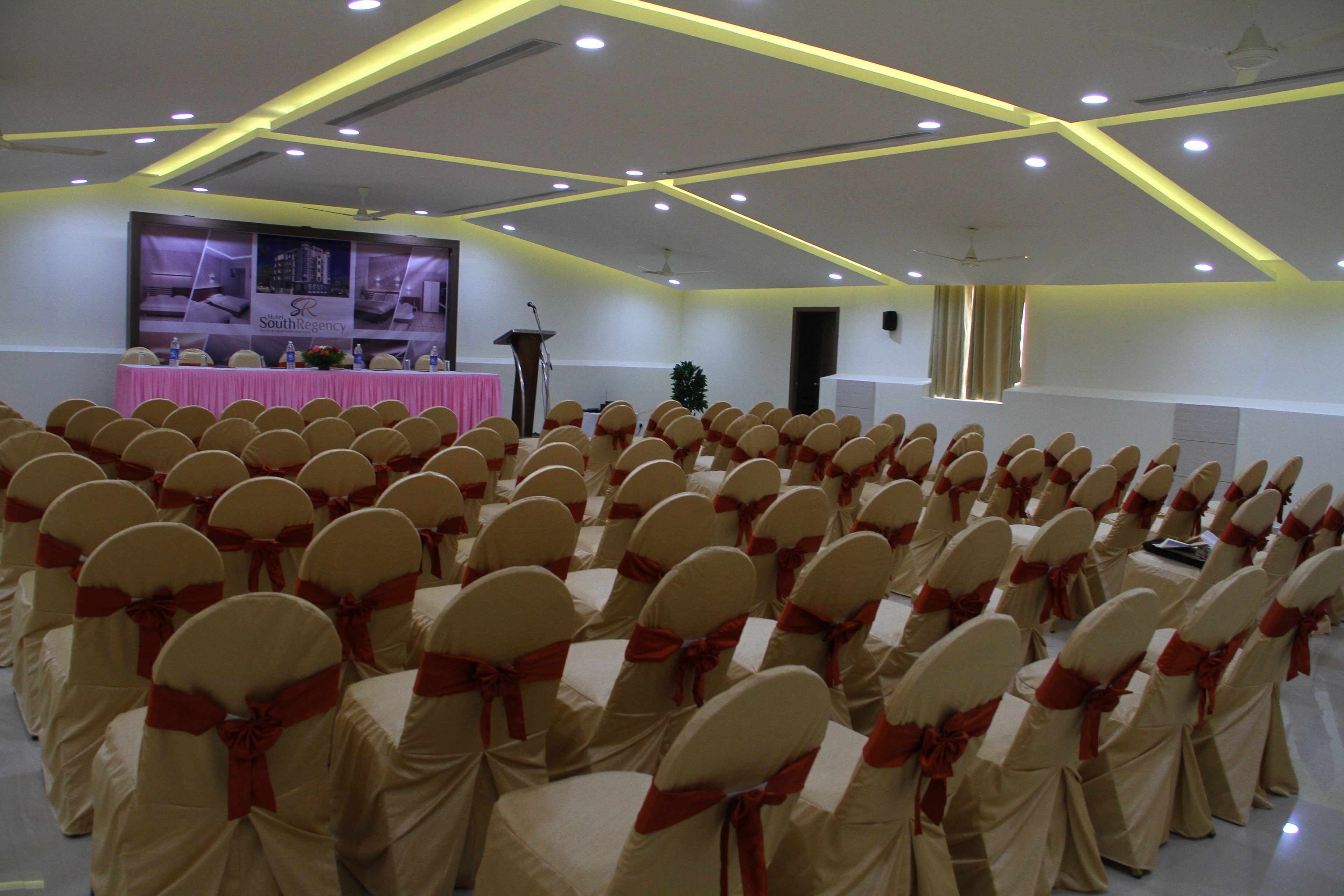Un salón de conferencias con filas de sillas cubiertas de tela color beige y listones rojos, un atril y un cartel en la pared.